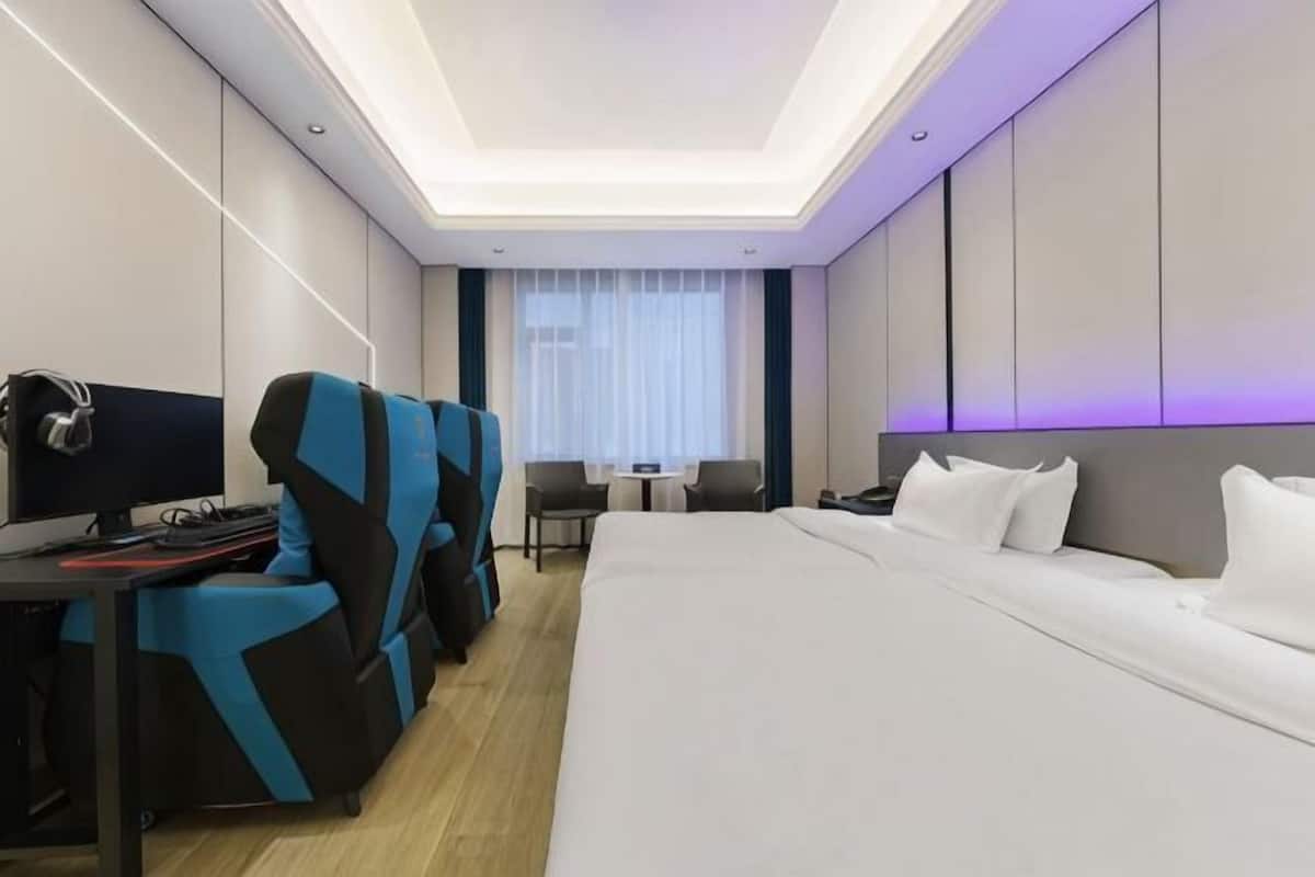 Ein modernes Hotelzimmer mit einem großen Bett, einem Schreibtisch mit Computer, einem blau-schwarzen Sessel und einem kleinen Tisch mit Stuhl.