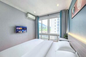 A hotel room with a bed, a mounted TV, a window with a view of buildings, and a balcony railing.
