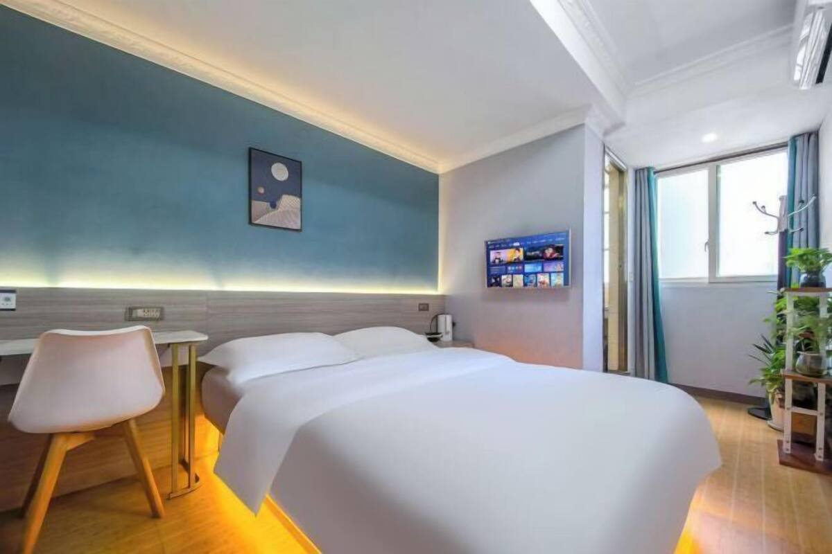 Habitación de hotel con cama, escritorio, silla, televisión instalada en la pared y ventana con cortinas.