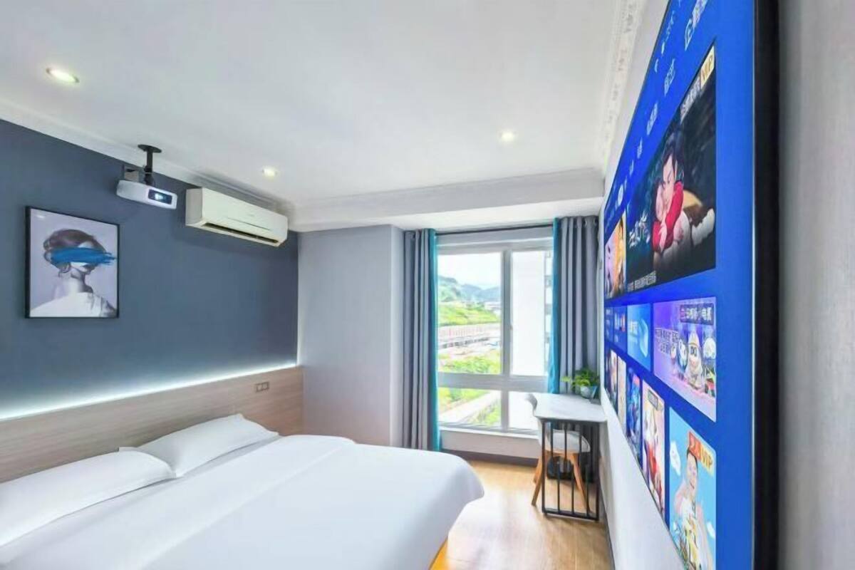 Una habitación de hotel con cama, escritorio, ventana grande, equipo de aire acondicionado y pared decorada con cuadros.