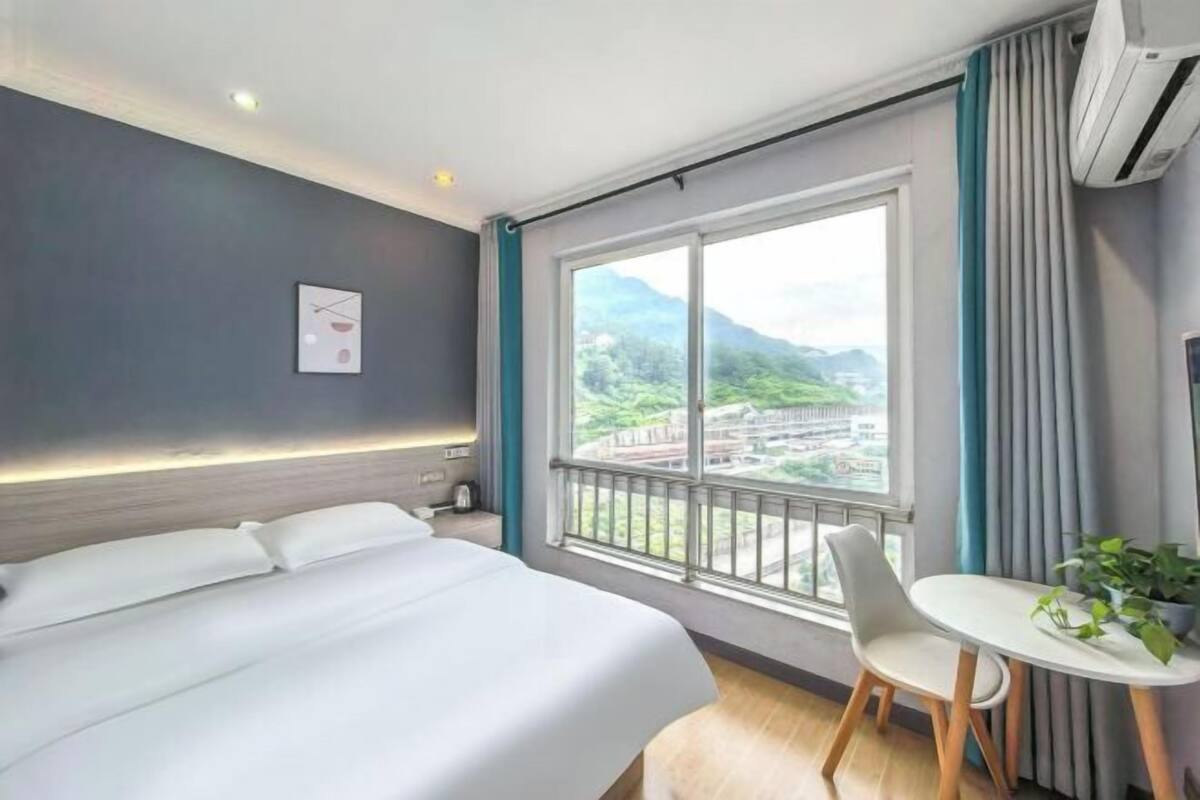 Habitación de hotel con una cama, una mesita con una silla, una ventana con vistas a montañas y un equipo de aire acondicionado.