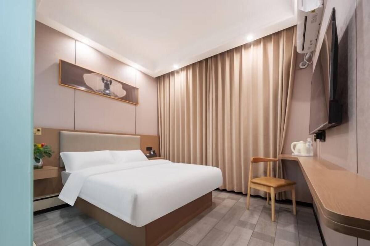 A hotel room with a bed, bedside table, chair, TV, curtains, and a painting on the wall.