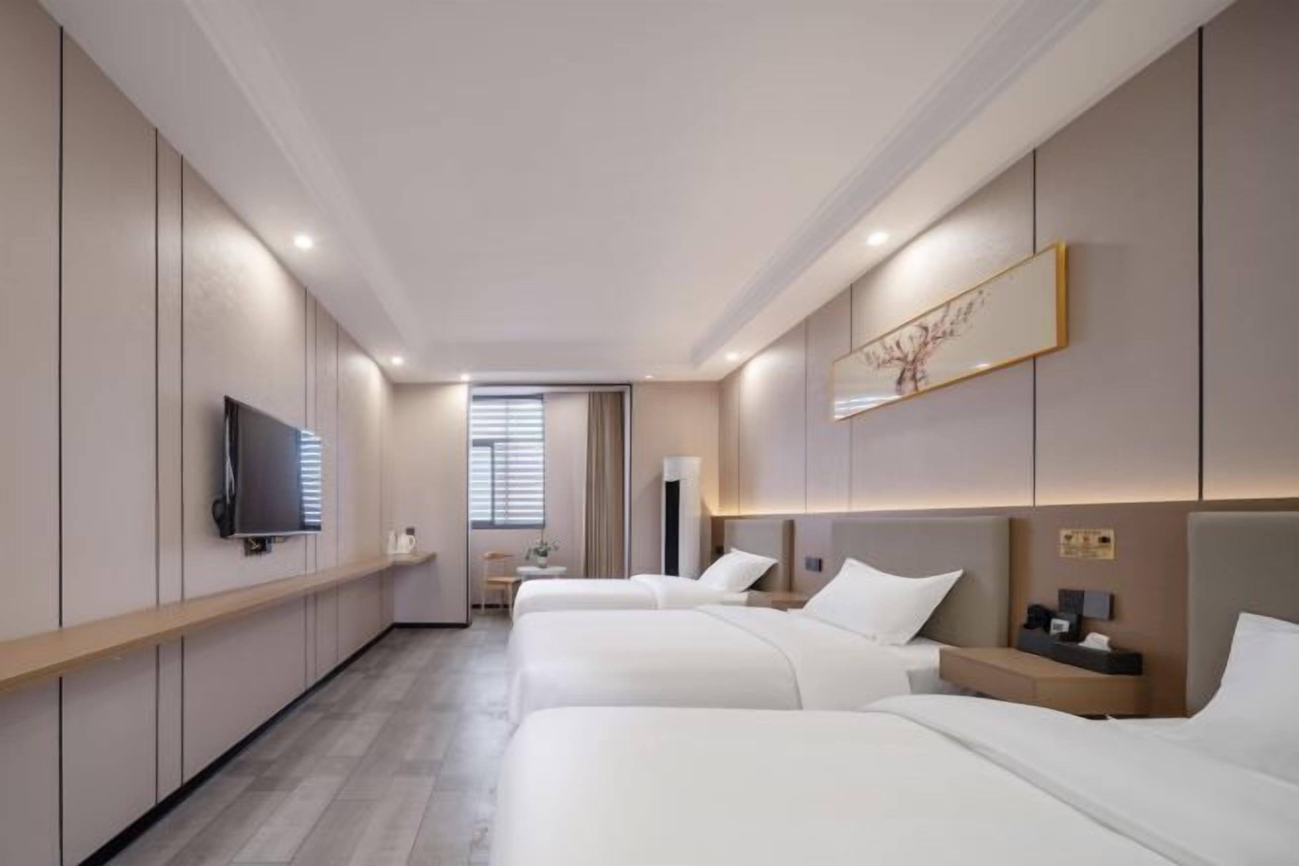 Habitación de hotel con tres camas, televisión, mesita de noche y ventana con persianas.