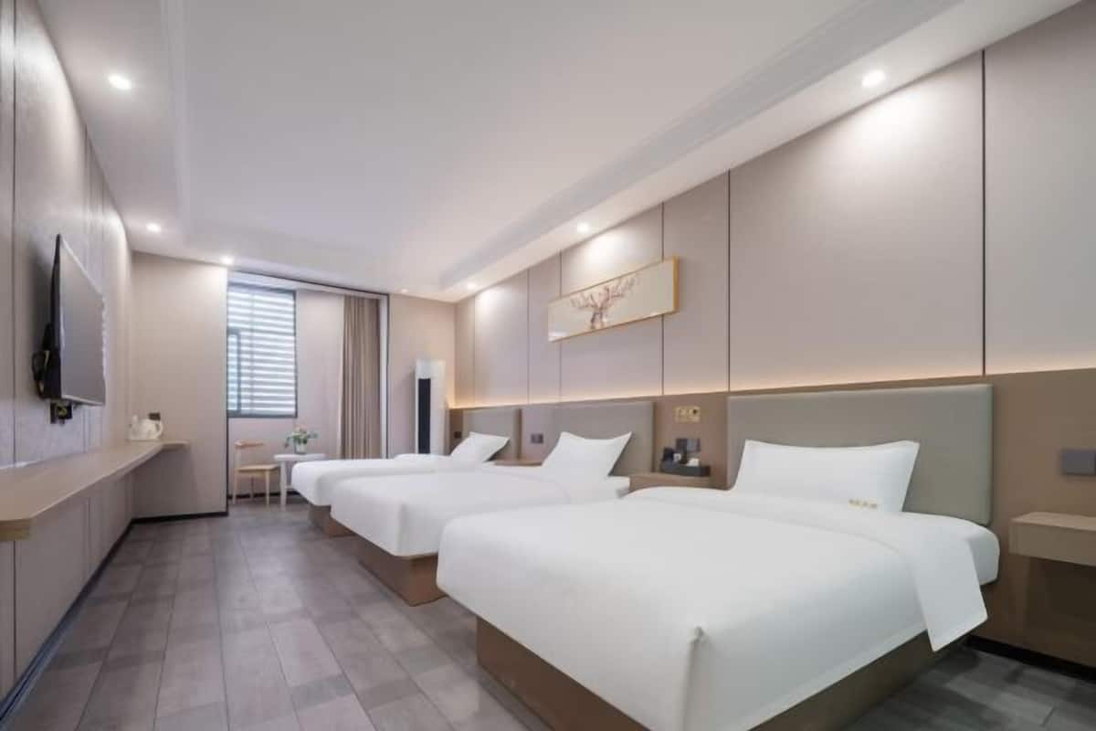 A hotel room with three beds, a TV, a small table, and a window with blinds.