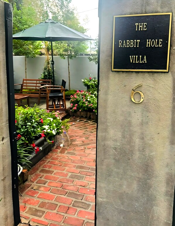 A brick pathway leading to a seating area with a bench and table, surrounded by potted plants and flowers. A sign reads "The Rabbit Hole Villa."