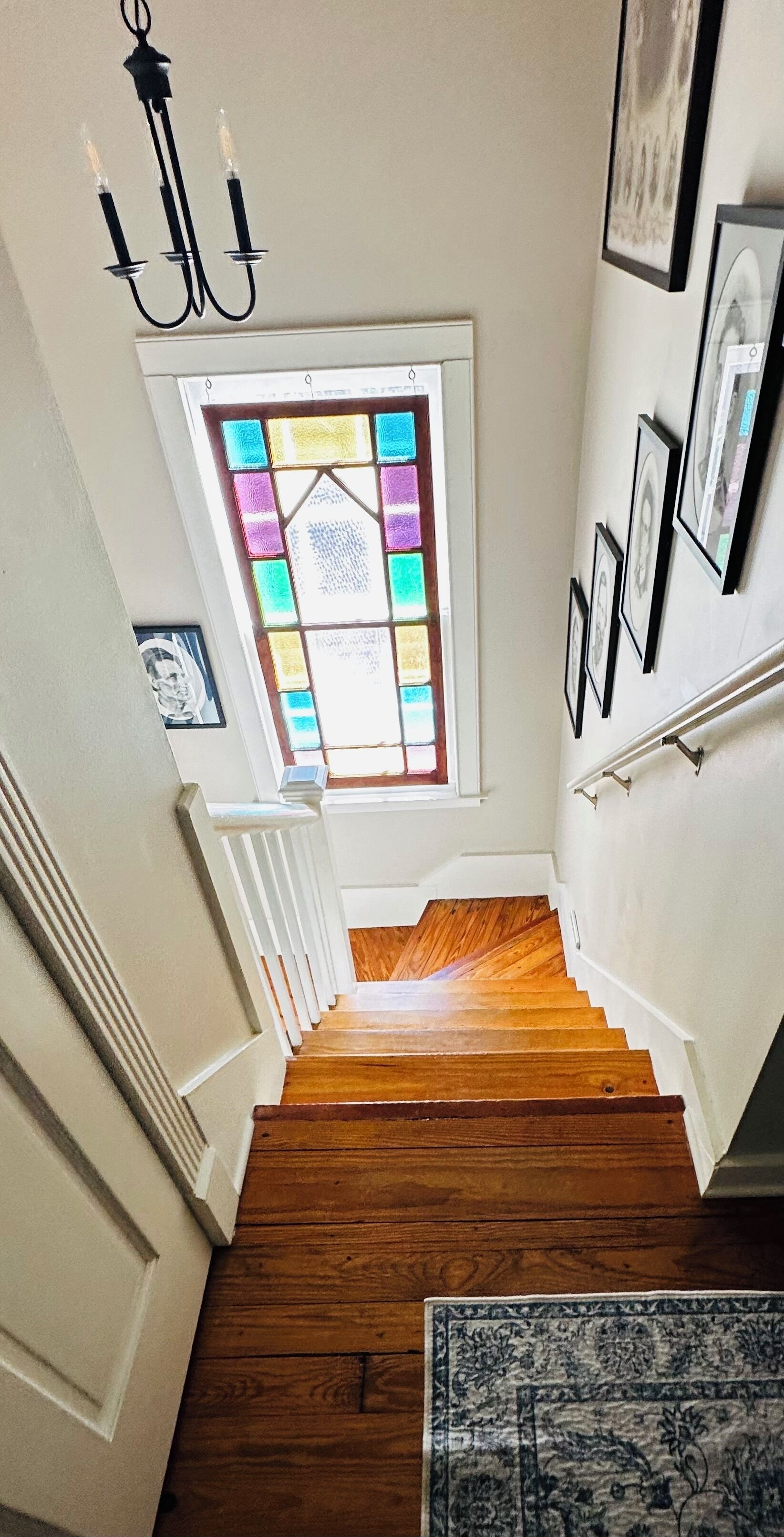 A wooden staircase with a stained glass window above, white walls, and framed pictures on the wall.