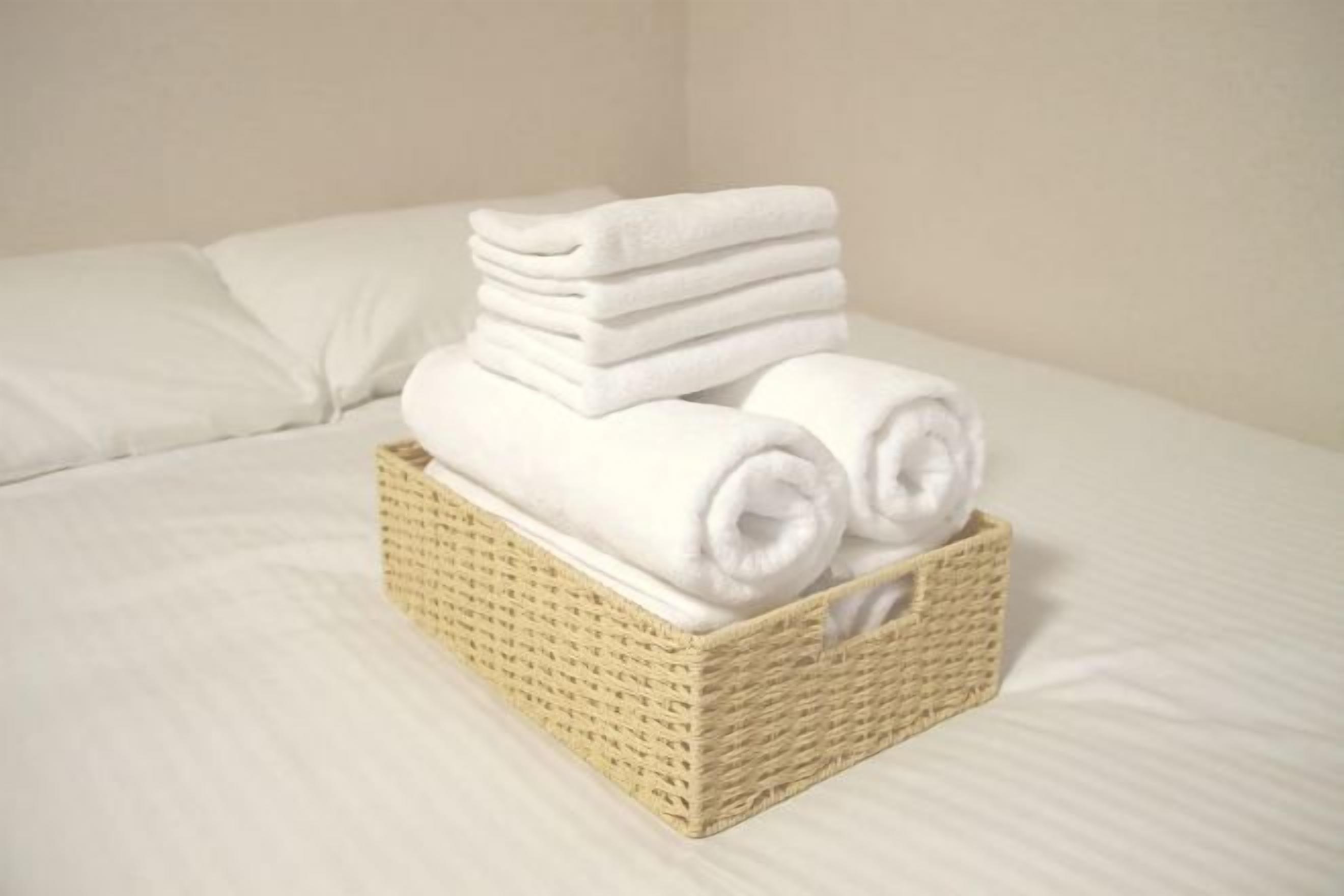 A neatly folded stack of white towels in a wicker basket on a bed with white bedding.