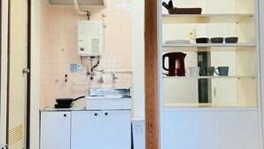 A kitchen with white cabinets, a stove, and a water heater. There is a shelf with kitchenware and a red kettle. - Kokoro no sato・Ren (Kyoto)