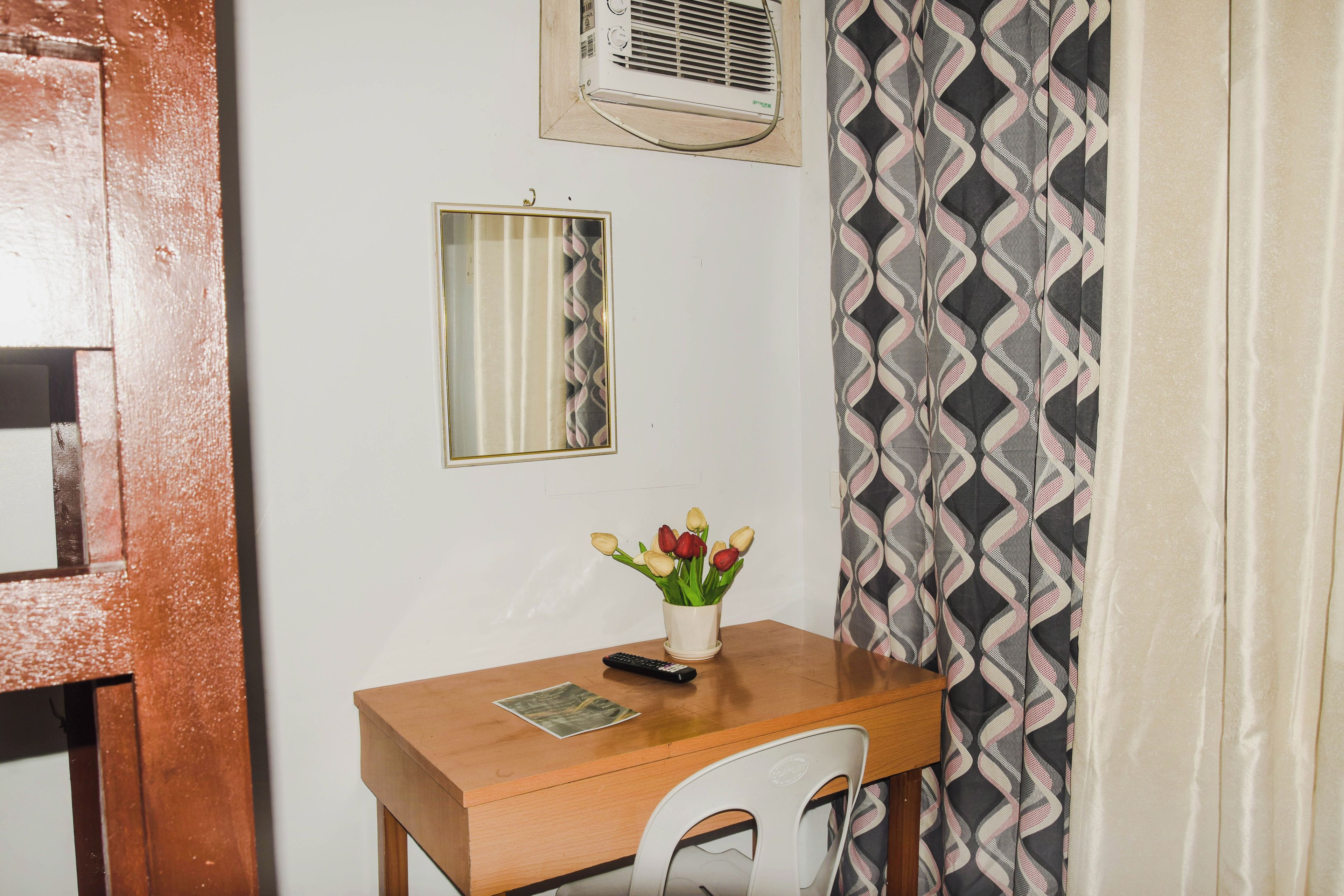 A small room with a wooden desk, a chair, a mirror, a wall-mounted air conditioner, and a patterned curtain.