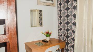 A small room with a wooden desk, a chair, a mirror, a wall-mounted air conditioner, and a patterned curtain.
