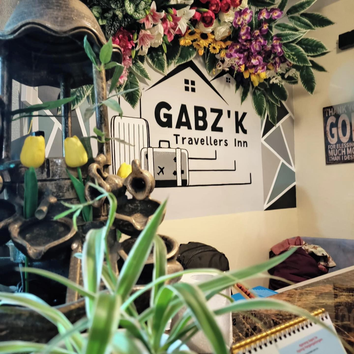 Una pared con el letrero 'GABZ'K Travellers Inn', rodeada de plantas decorativas y una escultura rústica.