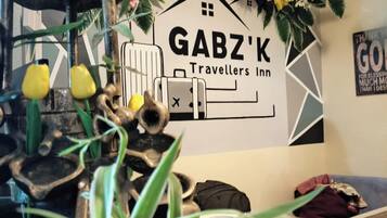 Una pared con el letrero 'GABZ'K Travellers Inn', rodeada de plantas decorativas y una escultura rústica.