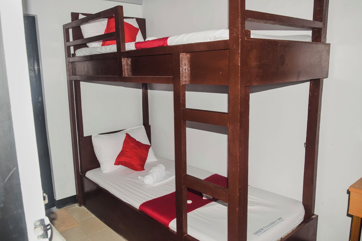 Uma cama beliche com roupa de cama branca e detalhes vermelhos, estrutura de madeira e uma pequena mesa de madeira ao lado.