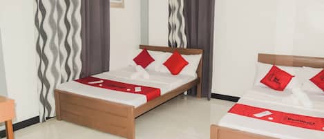 A hotel room with two beds, each with a wooden frame and white bedding. There are red pillows and a red blanket on one bed, and a red towel on the other. The room has a tiled floor and curtains with a wavy pattern.