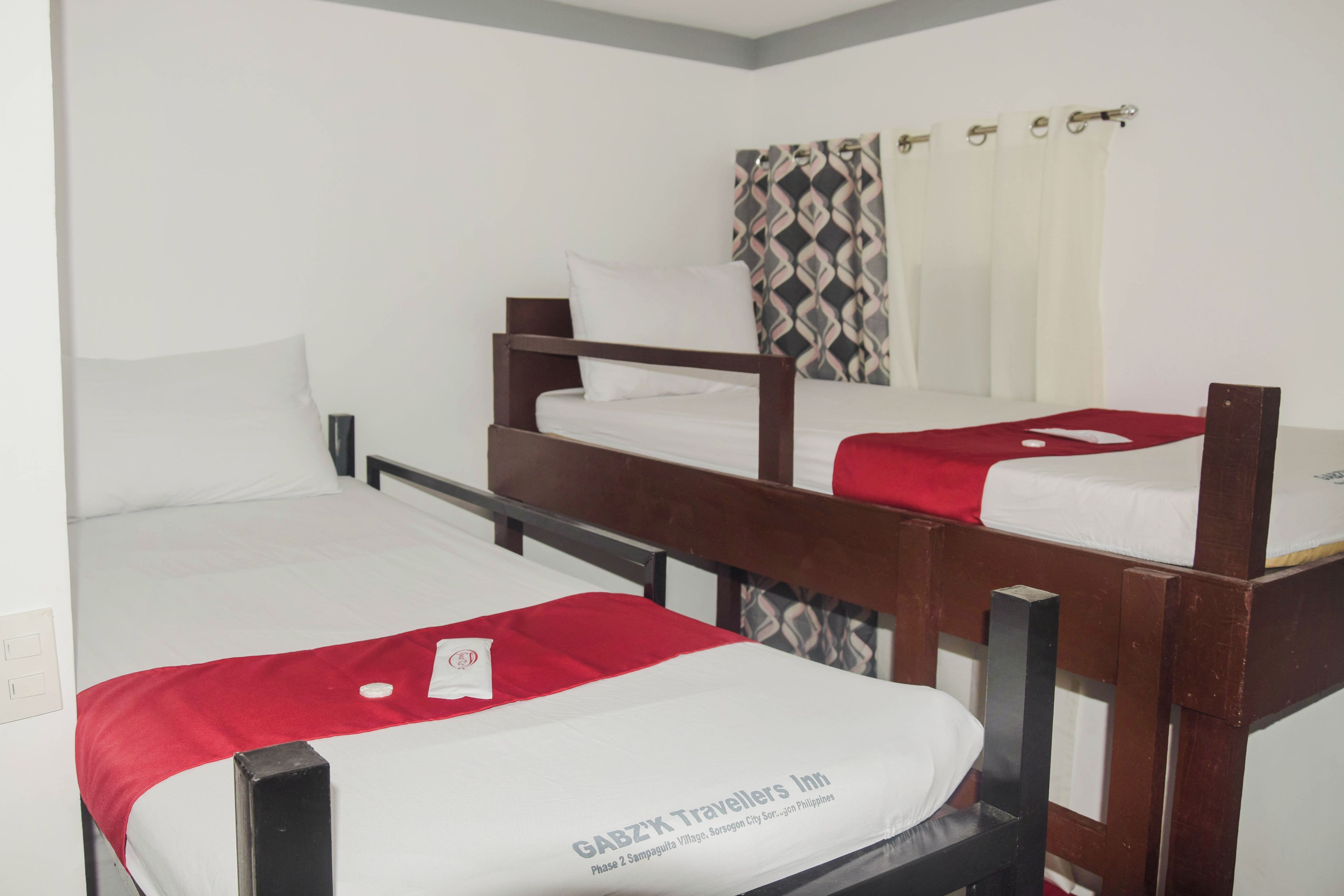 Quarto com três beliches, cada um com colchão branco e cobertor vermelho. Os beliches possuem estrutura de madeira e estão dispostos em linha. Há uma cortina estampada na janela atrás do beliche do meio.