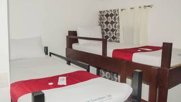Quarto com três beliches, cada um com colchão branco e cobertor vermelho. Os beliches possuem estrutura de madeira e estão dispostos em linha. Há uma cortina estampada na janela atrás do beliche do meio.