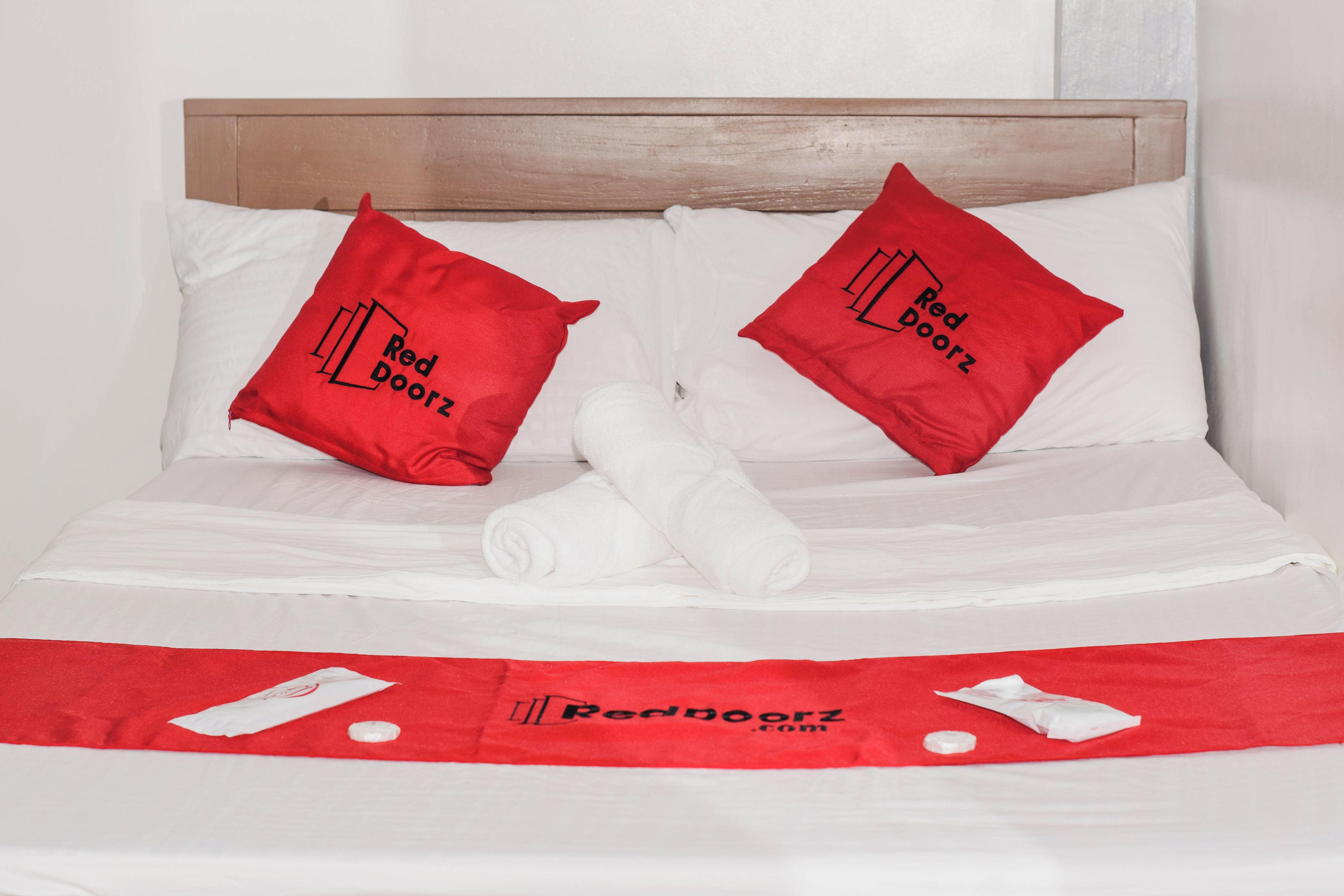 Cama bem arrumada com duas almofadas vermelhas e uma toalha branca, uma toalha vermelha sobre a cama, e um edredom vermelho com o logotipo Red Doorz.