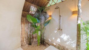 Un área de ducha con techo de bambú, cabezal de ducha en la pared y una planta grande.