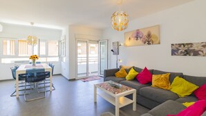 Un salon moderne avec un canapé gris, des coussins colorés, une table basse blanche et un coin repas comprenant une table et des chaises.