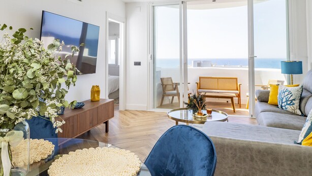 A modern living room with a glass coffee table, a blue chair, a sofa with yellow and blue cushions, and a large TV on the wall. - Cubo's Vistalmar Calahonda Apartment / beach access / terrace / communal pool / free wifi (Mijas Costa)