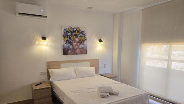 A bedroom with a bed, bedside tables, an air conditioner, a painting, and a window with blinds. - Hotel Key Reig (Almussafes)