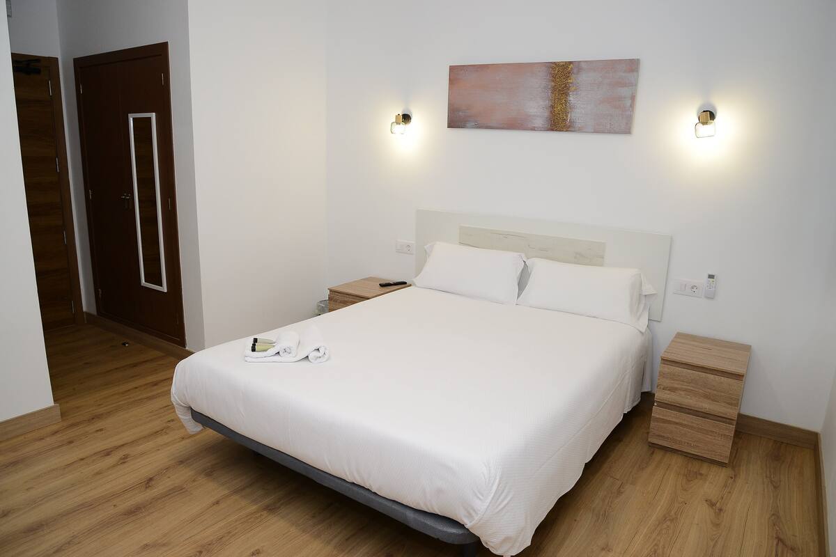 A neatly made bed with white linens, a wooden nightstand with a lamp, and a wooden drawer unit beside it.
