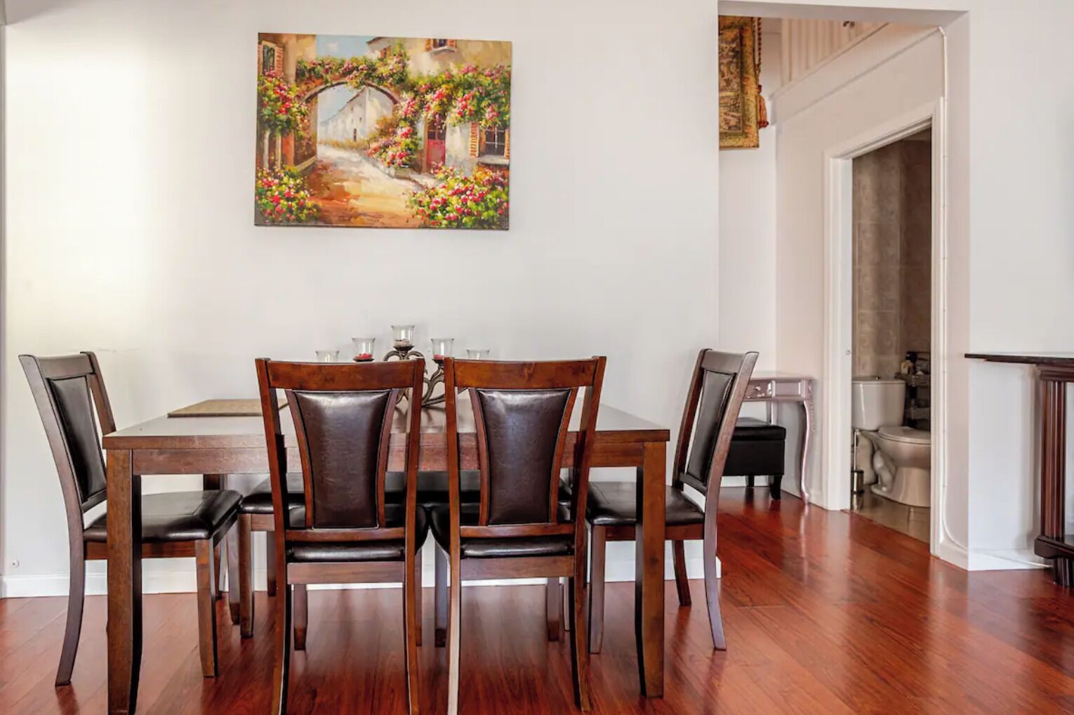 A dining room with a wooden table and chairs, a painting on the wall, and a view into the adjacent bathroom.