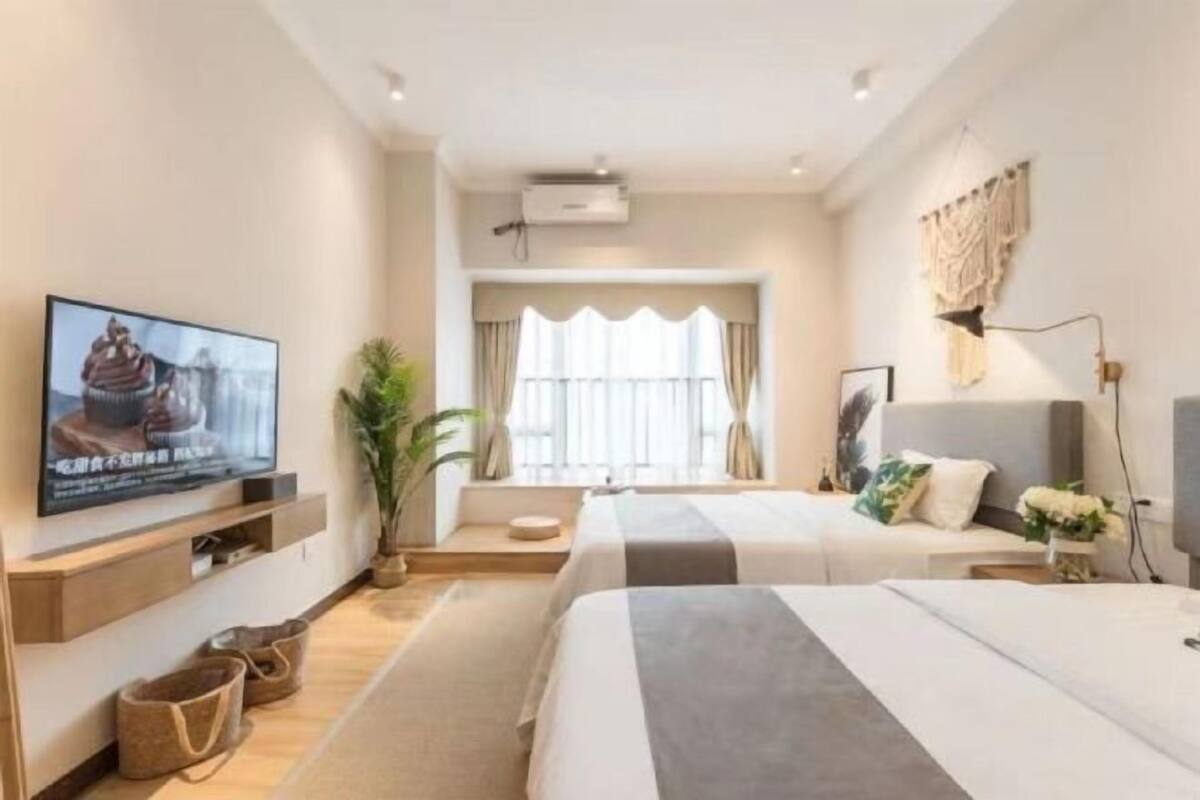 Quarto de hotel com duas camas, televisão, janela com cortinas, planta e ar-condicionado fixado na parede.