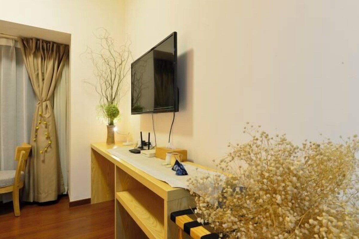 A room with a wooden desk, a flat-screen TV mounted on the wall, a chair, and a window with curtains.