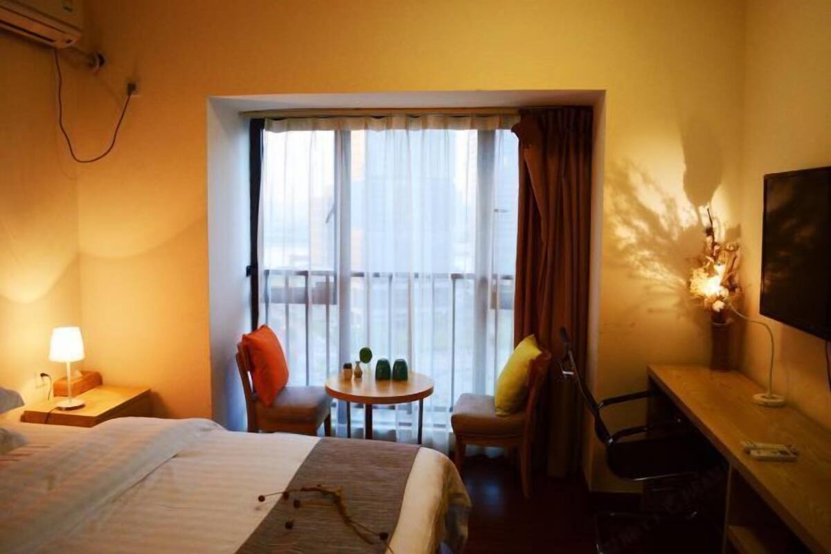 Quarto de hotel com cama, escrivaninha, cadeira, mesinha e janela com cortinas.