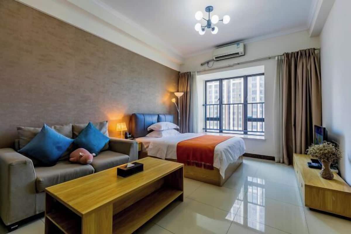 Quarto de hotel com sofá, cama, mesa de centro de madeira, abajur e janela com cortinas.