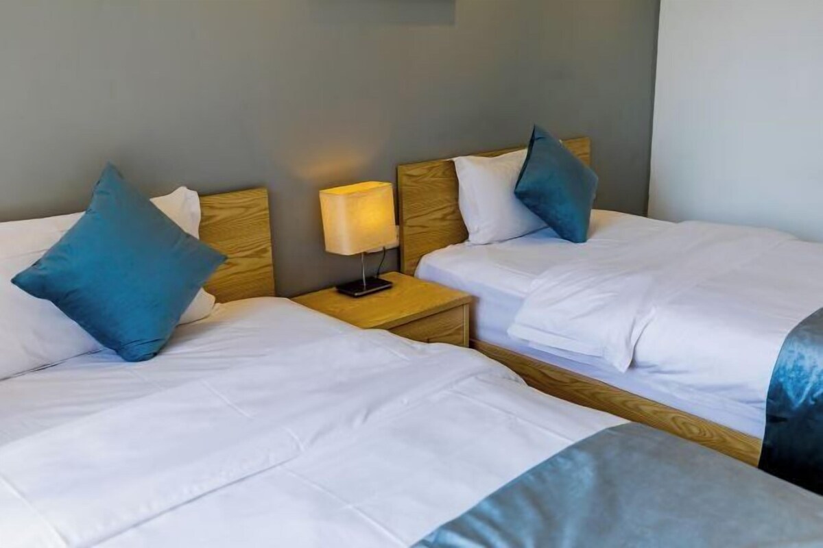 A hotel room with two beds, each with white bedding and blue pillows. There is a bedside table with a lamp and a smartphone on it.