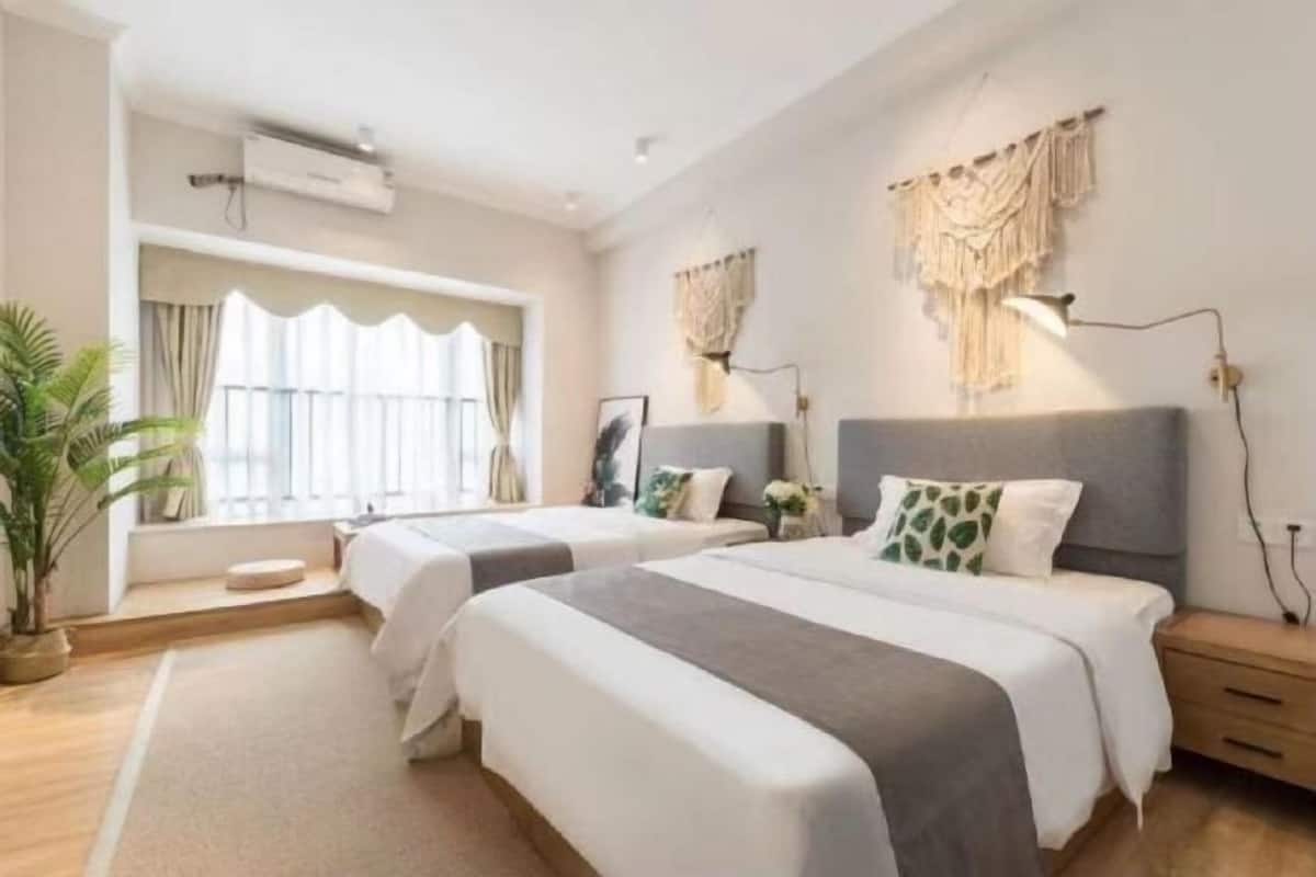 Quarto de hotel com duas camas, uma janela grande com cortinas, uma planta em vaso e um ar condicionado fixado na parede.