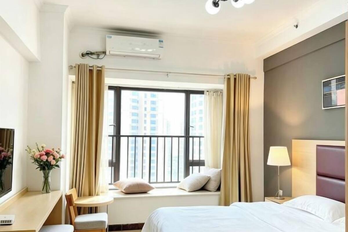Quarto de hotel com cama, escrivaninha, cadeira, janela com cortinas, ar condicionado e abajur.