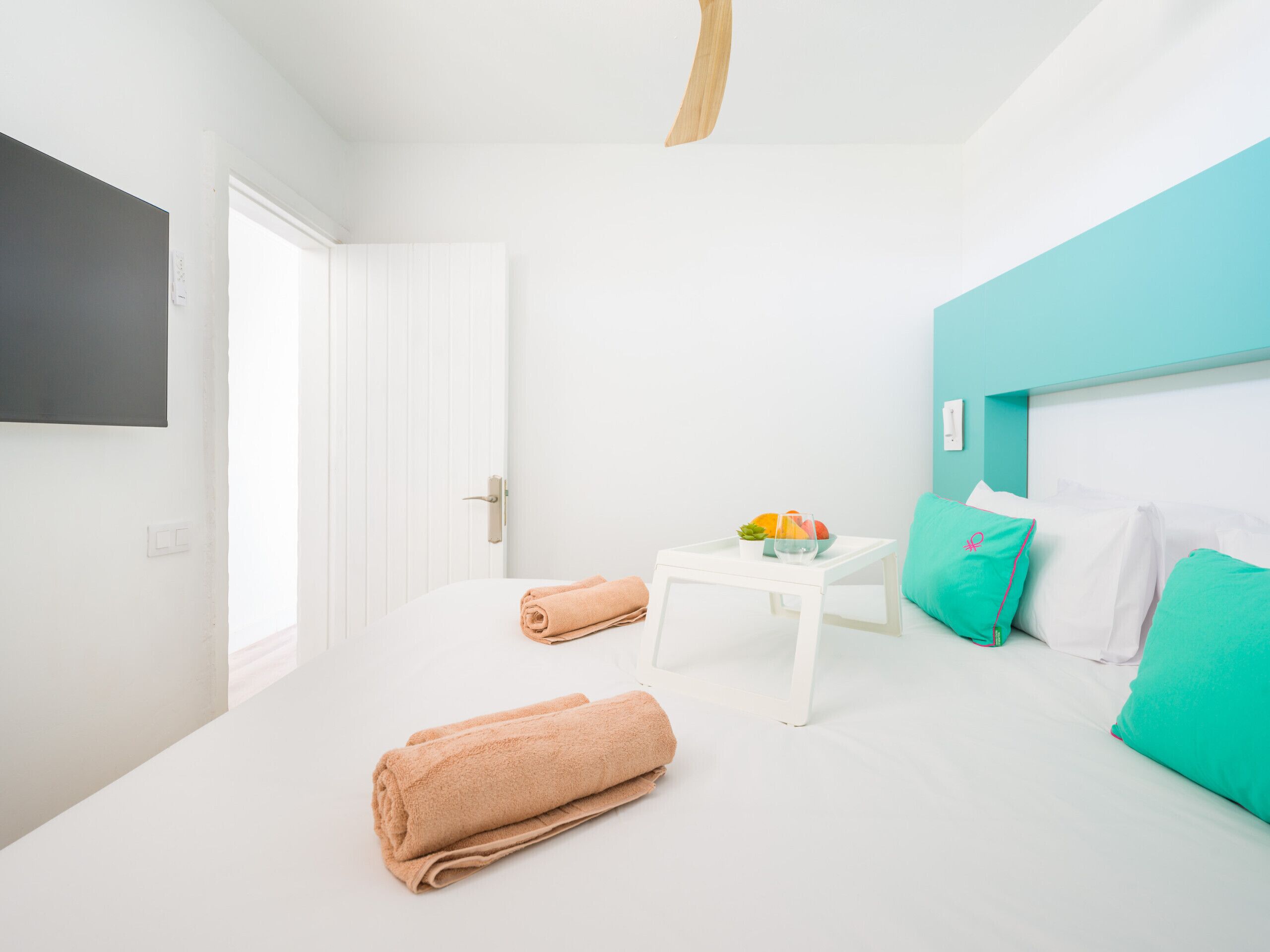 Habitació d'hotel moderna amb un llit, una taula petita amb fruita i un got, una tovallola estovada i una paret d'accent color turquesa.