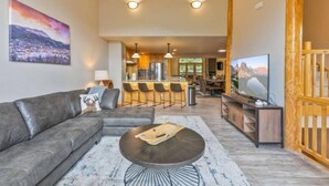 A modern living room with a gray sofa, a round coffee table, and a TV on a wooden stand. There is a kitchen area with bar stools and a dining table in the background. - 5 Bedroom Luxury Townhome Near Snowflake Lift in Breckenridge - Walk to Lift! (Breckenridge)