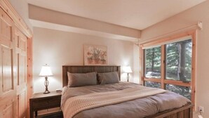 A bedroom with a wooden bed frame, two bedside tables with lamps, a large window with a view of trees, and a painting on the wall. - 5 Bedroom Luxury Townhome Near Snowflake Lift in Breckenridge - Walk to Lift! (Breckenridge)