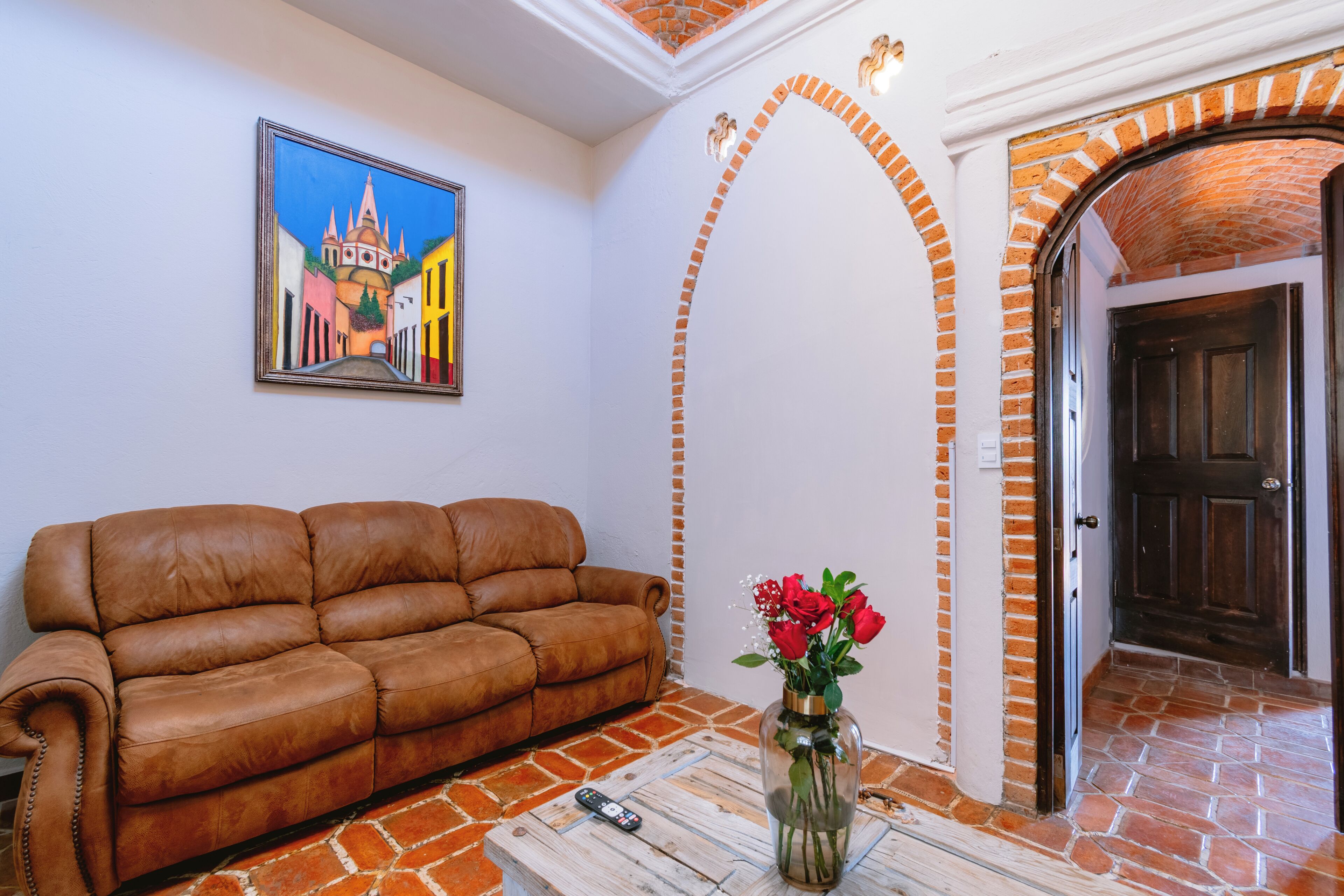 Un saló amb un sofà de cuir marró, un ram de flors vermelles, un comandament a distància i un quadre d'un edifici a la paret.