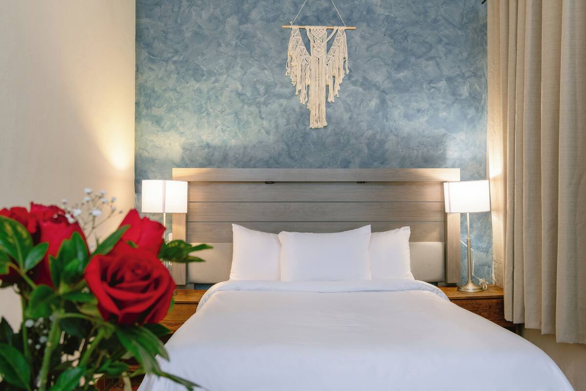 Une chambre d’hôtel avec un grand lit, du linge de lit blanc, une décoration murale en macramé et deux lampes de chevet.