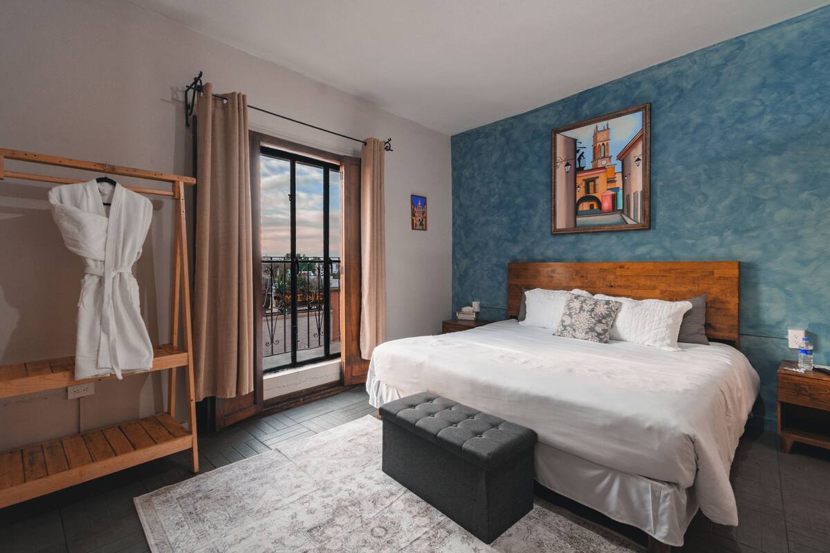 Une chambre avec un grand lit, une tête de lit en bois, une table de chevet, un banc, un porte-manteau et une vue sur l’extérieur grâce à une porte coulissante.