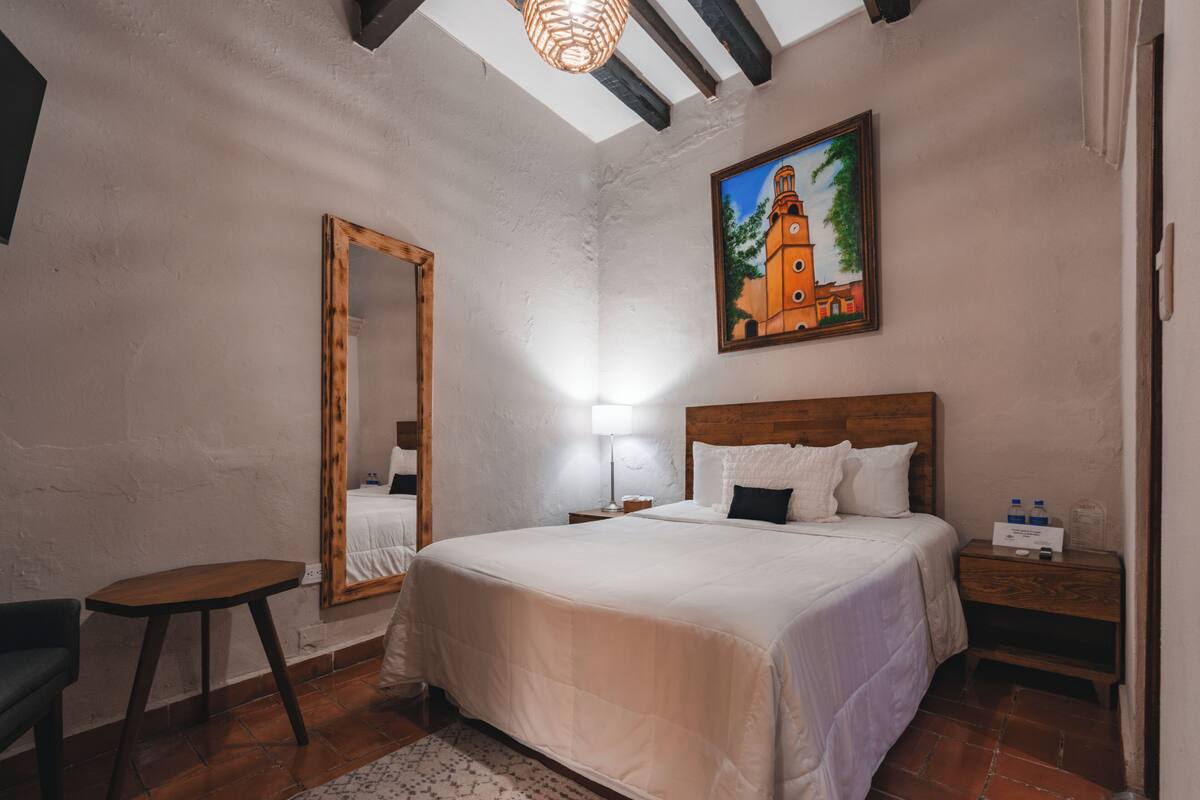 Une chambre à coucher avec un grand lit, une table de chevet en bois, une petite table, un miroir et un tableau représentant une église accroché au mur.
