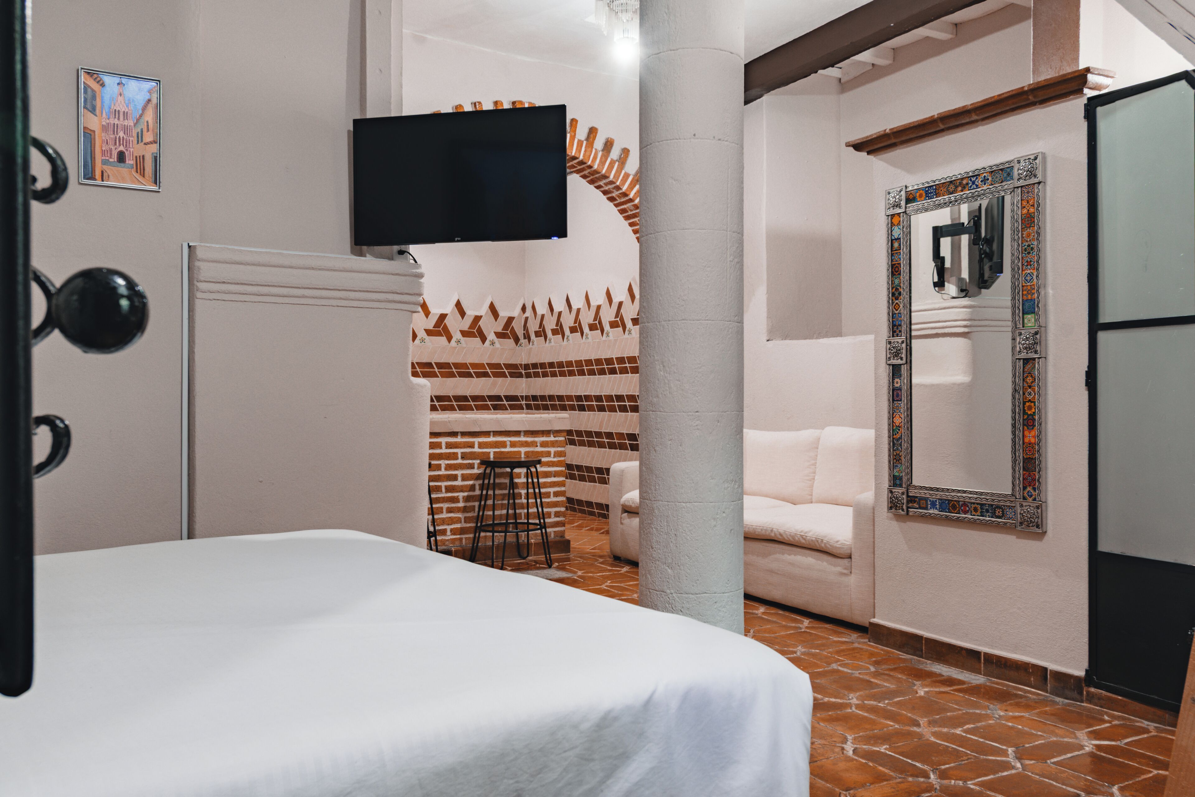 Habitación de hotel con cama, televisor montado en la pared, sofá blanco y espejo estampado.