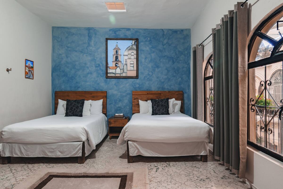 Une chambre d’hôtel avec deux lits, un mur d’accent bleu, un tableau encadré et une fenêtre donnant sur l’extérieur.