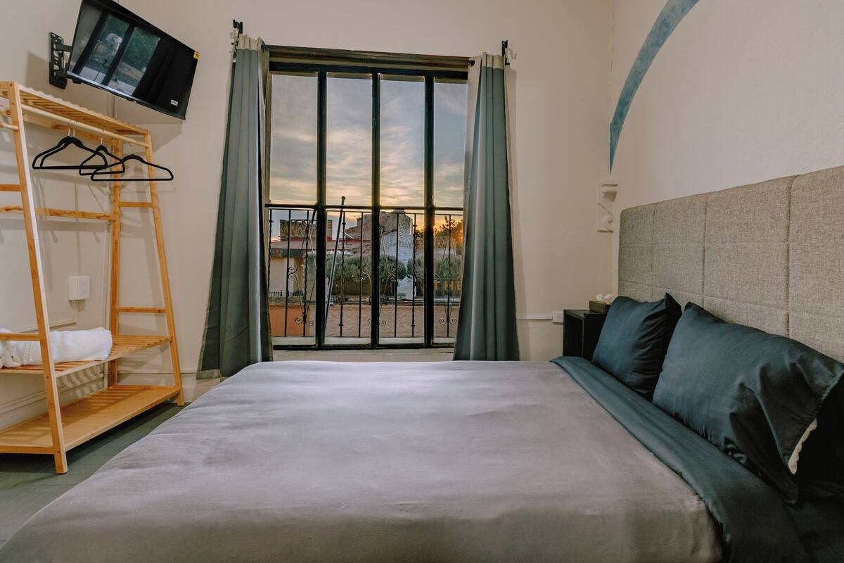 Une chambre d’hôtel avec un grand lit, un téléviseur fixé au mur, une étagère en bois et une vue sur l’extérieur par une fenêtre munie de rideaux.