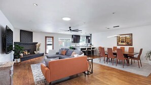 Uma sala de estar ampla com lareira, um sofá, um espaço de jantar com mesa e cadeiras, e uma cozinha ao fundo.