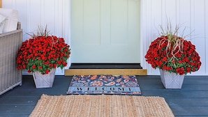 A front door with two potted plants, a welcome mat, and a wicker chair to the left. - Enchanting 2 Bedroom Cottage, The Fox Den, 258 Acres, Farmville, VA (Rice)