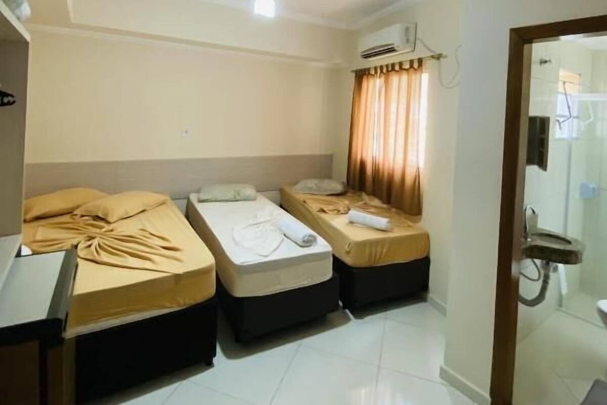 Habitación de hotel con dos camas individuales, un ventilador de techo, un equipo de aire acondicionado y un baño visible a través de un vidrio.