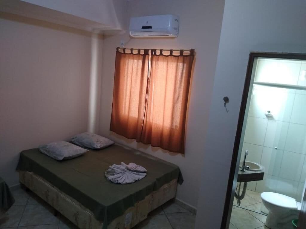 A small room with a bed, a window with curtains, a wall-mounted air conditioner, and a view of a bathroom with a toilet and sink.