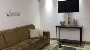 A living room with a brown sofa, a coffee table with decorative items, a flat-screen TV on a stand, and a patterned rug. - Basilica Palace Hotel (Aparecida)