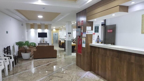 A reception area with a wooden reception desk, a seating area with a sofa and chairs, and a potted plant. - Basilica Palace Hotel (Aparecida)